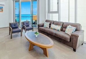 A modern living room with a brown leather sofa, two armchairs, and a round coffee table. A large window offers a view of the ocean. - New Modern Exclusive Oceanview Condo (Noord)