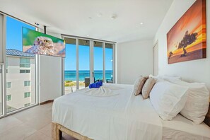 A bedroom with a large bed, a TV, and a painting of a sunset. - New Modern Exclusive Oceanview Condo (Noord)