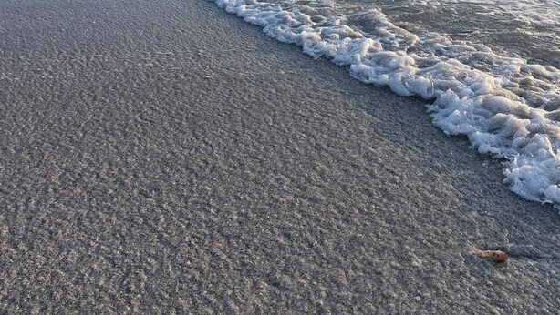 Uma praia com ondas suaves, céu claro e um píer ao longe.