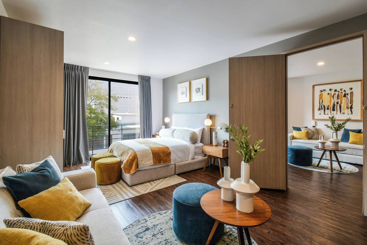 A modern hotel room with a large bed, a sofa, a coffee table, and a view of the outdoors.