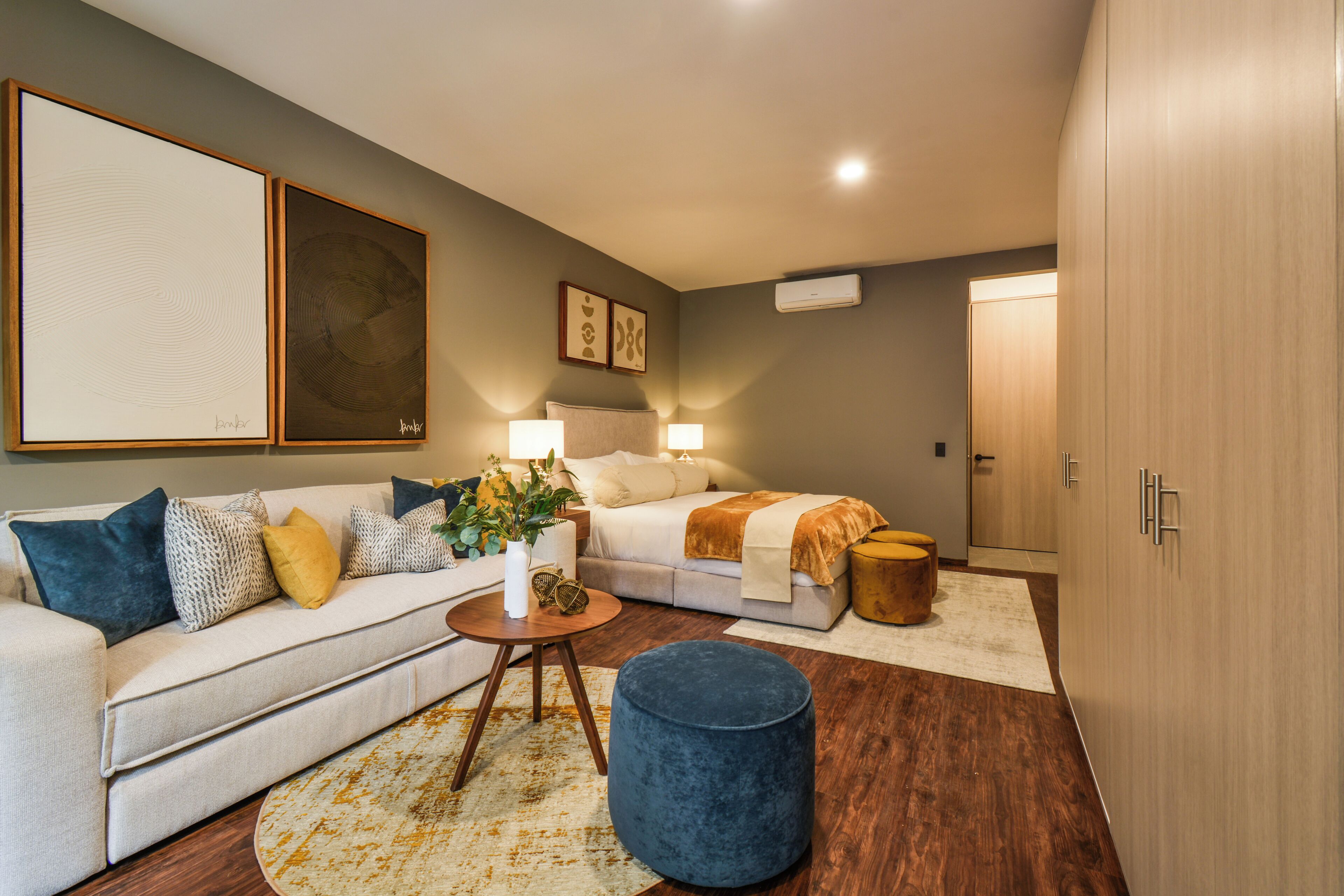 Un salón moderno con sofá, una mesita redonda y una cama con cabecero. Se observan unidades de aire acondicionado fijadas a la pared y cuadros enmarcados sobre el sofá.