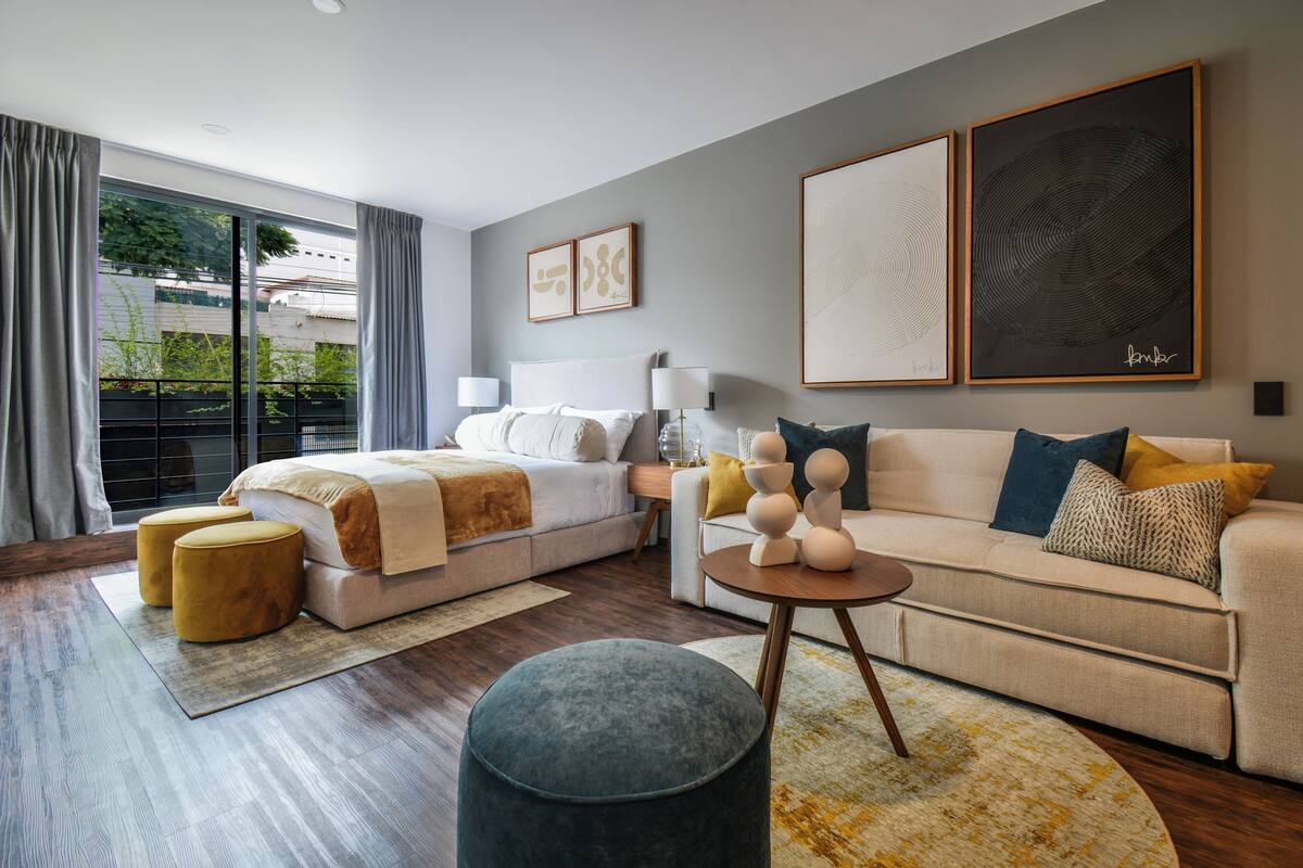 A modern hotel room with a large bed, a sofa, two ottomans, and a coffee table. There are two framed artworks on the wall and a window with curtains.