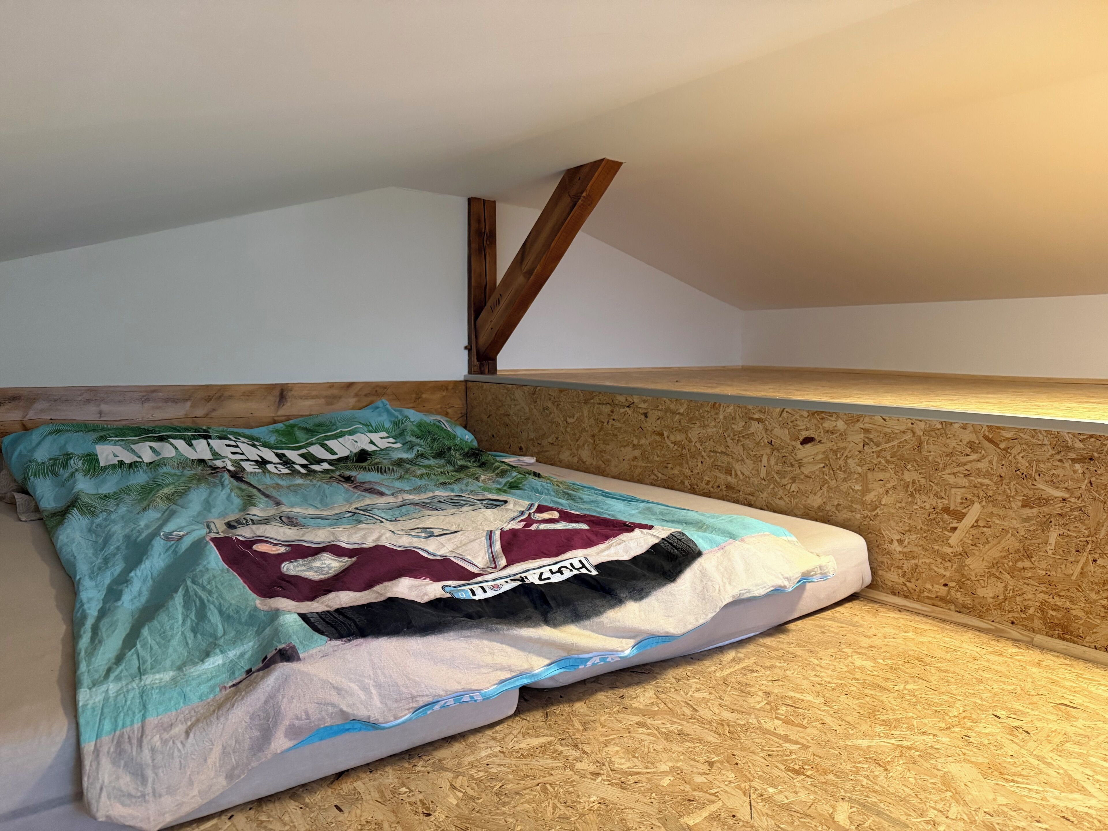 Une petite pièce avec un plafond en pente, où se trouve un lit adossé à un mur en bois, agrémentée d’une couverture colorée.