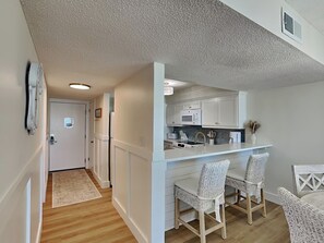 A compact kitchen with white cabinets, a microwave, and a sink. There are two wicker chairs and a small dining table. - Dunes of Panama #C503 | Gulf Front + Amenities (Panama City Beach)