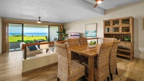 A dining area with a wooden table and wicker chairs, a ceiling fan, and a view of the ocean through large windows. - Puu Poa 102 - Private and Serene 2/2 with ocean views (Princeville)
