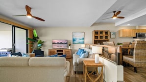 A modern living room with a sofa, coffee table, and a dining area with wicker furniture. There are ceiling fans, a television, and wooden cabinets. - Puu Poa 102 - Private and Serene 2/2 with ocean views (Princeville)
