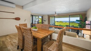 A dining area with a wooden table and wicker chairs, a ceiling fan, and a view of the ocean through large windows. - Puu Poa 102 - Private and Serene 2/2 with ocean views (Princeville)