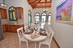 A dining area with a round table, white chairs, and a view of the outdoors through large windows. - Villa Poseidon (Corfu)
