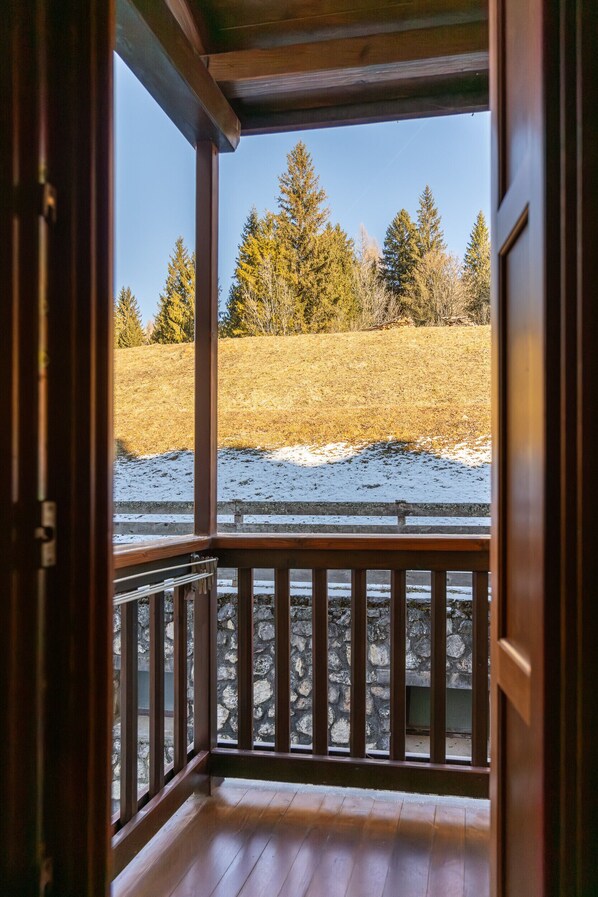 Un balcon en bois, doté de balustrades en pierre, surplombe un champ verdoyant parsemé d’arbres et recouvert de neige.