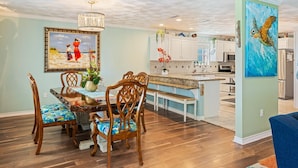 A dining area with a wooden table and chairs, a chandelier, and a painting of a beach scene. - Casa Bella - 4 bedroom, Gulf View, Cargo Lift, Outdoor Bar, Fenced Yard, (Crystal Beach)