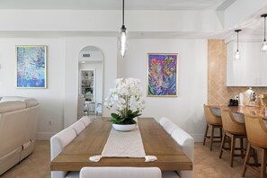 A dining area with a wooden table, white chairs, a white sofa, and a vase of white orchids. There are two framed paintings on the walls and a kitchen area with white cabinets and a tiled backsplash. - Turquoise Place 2102C by Liquid Life (Orange Beach)