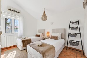 Ein Hotelzimmer mit zwei Betten, einer Pendelleuchte, einer Leiter mit Körben und einem Fenster mit Vorhängen.