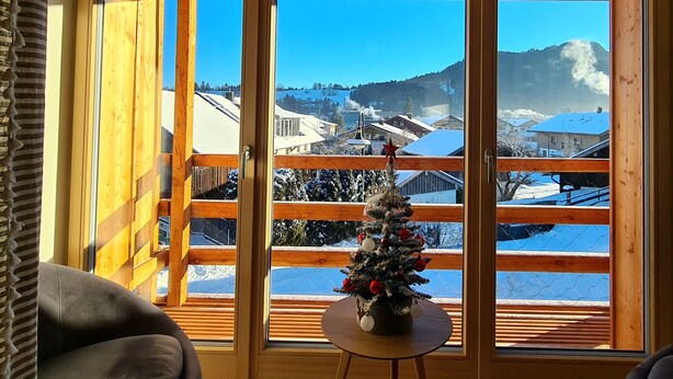 Una habitación con un amplio ventanal, una mesita con un árbol de Navidad y vistas a un pueblo cubierto de nieve.