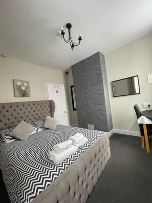 Une chambre à coucher moderne avec une tête de lit capitonnée, un grand lit recouvert d’une couverture à motifs zigzag noir et blanc, un banc rembourré de couleur grise, un bureau avec une chaise, un miroir et un luminaire fixé au mur.