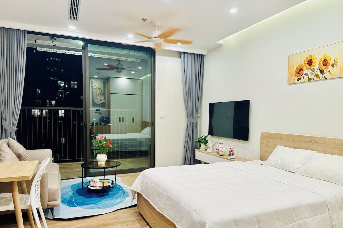 Ein modernes Hotelzimmer mit einem großen Bett, einem an der Wand montierten Fernseher, einem Balkon mit Blick auf die nächtliche Stadt und einer Essecke mit Tisch und Stühlen.