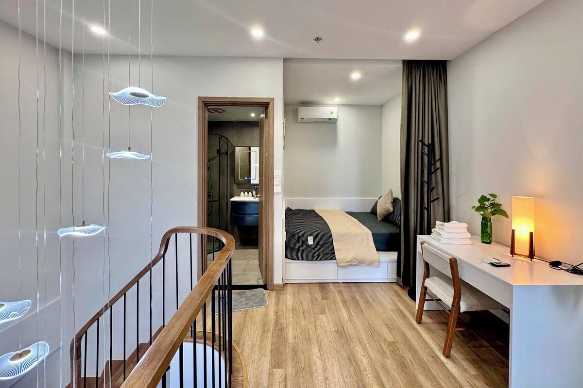 Ein modernes Hotelzimmer mit einer Holztreppe, einem Schreibtisch mit Lampe, einem Bett und einem durch eine offene Tür einsehbarer Badezimmer.