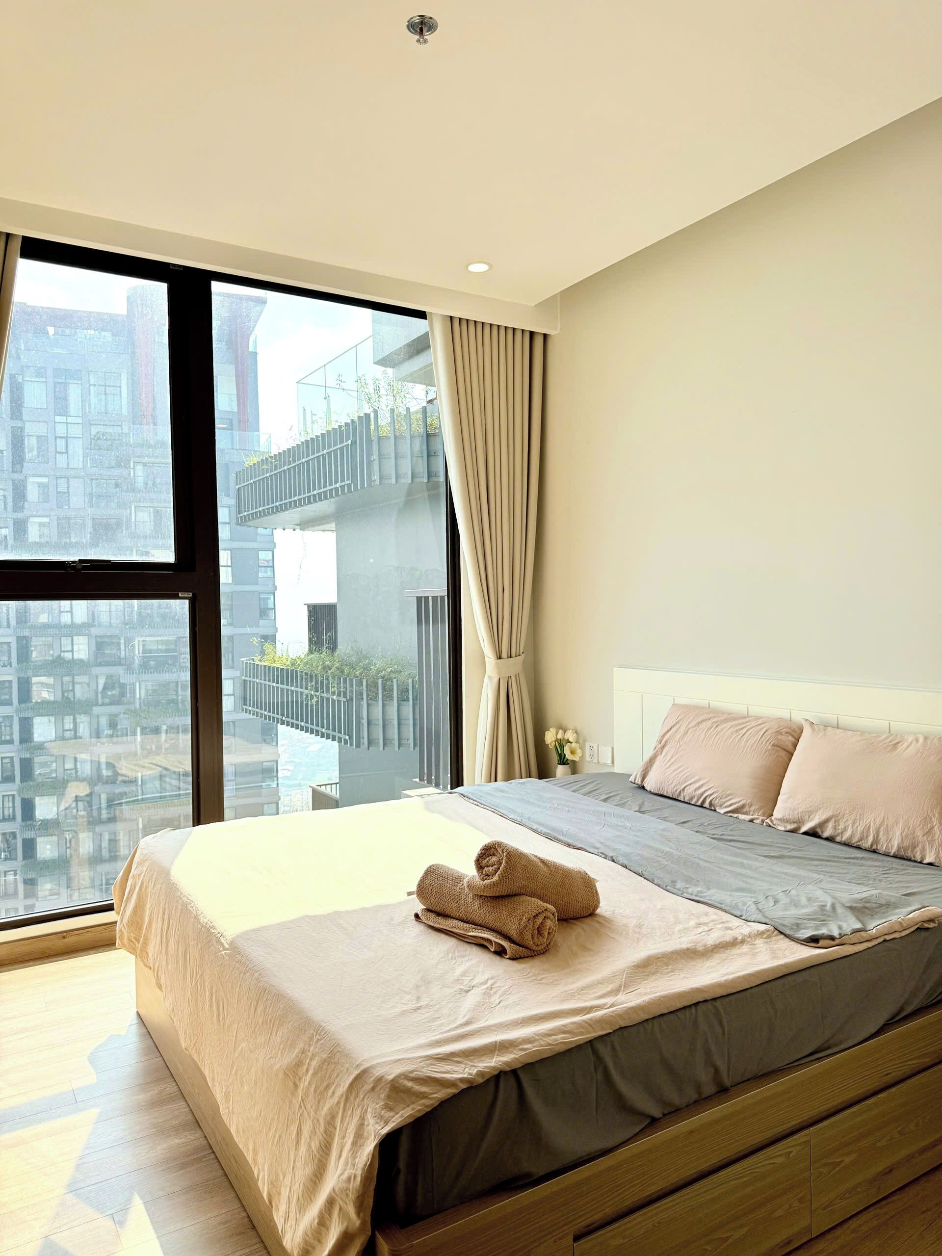Ein modernes Schlafzimmer mit einem großen Fenster, einem Bett mit beige farbenen Textilien und Blick auf eine Stadtlandschaft.