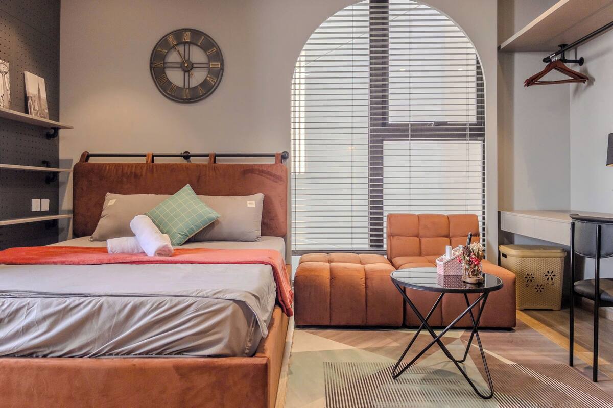 Ein modernes Schlafzimmer mit Bett, rundem Couchtisch, Sofa und großem Fenster mit Jalousien.