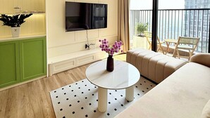 A modern hotel room with a large window, a flat-screen TV, a round coffee table with a vase of flowers, a sofa, and a green cabinet.