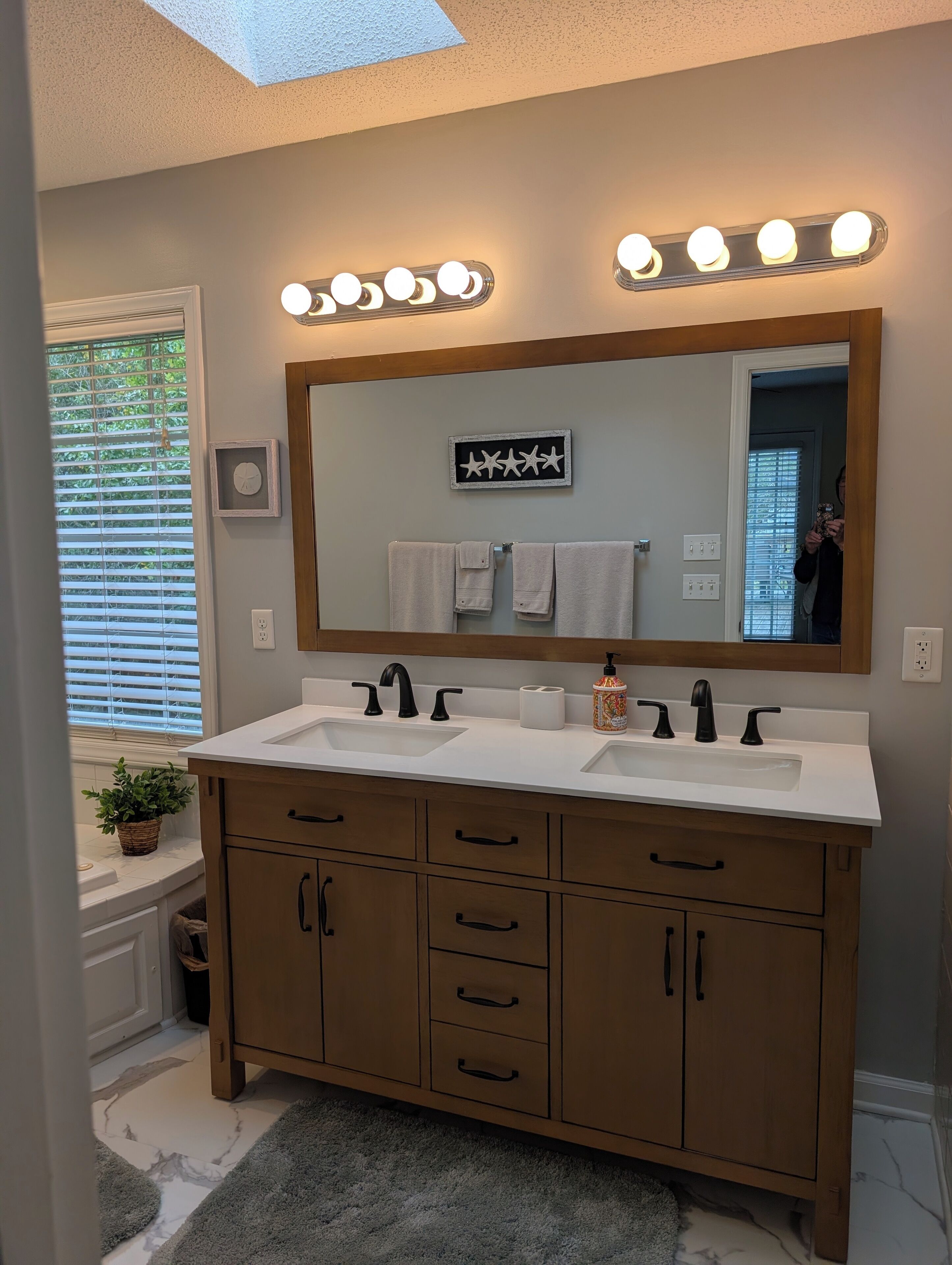 A bathroom with a double sink vanity, a large mirror with lights, and a potted plant.