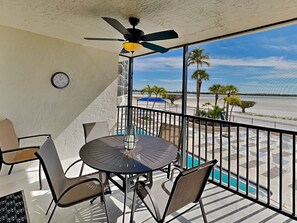 A balcony with a table and chairs, a ceiling fan, and a view of the beach and palm trees. - Carlos Pointe 121 | Stylish Retreat  w/Gulf Views, Balcony + Community Pool (Fort Myers Beach)