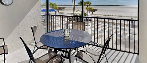 A balcony with a round table and chairs, a clock on the wall, and a view of a beach with palm trees.