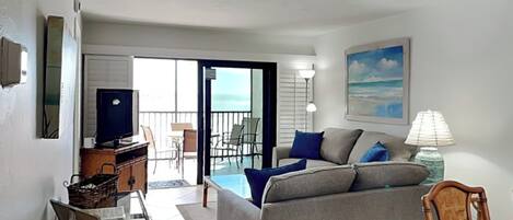 A living room with a sofa, a TV, a lamp, and a view of the ocean through the balcony doors.