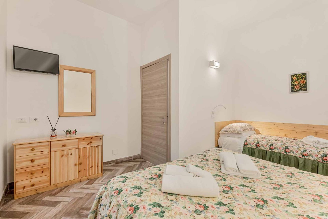 Ein Hotelzimmer mit einem hölzernen Schminktisch, einem an der Wand montierten Fernseher, einem Bett mit floralem Bettzeug und einer kleinen Wandleuchte.