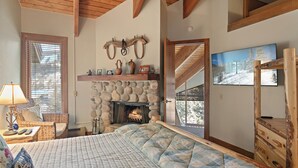 Ein gemütliches Schlafzimmer mit Kamin aus Stein, einem Bett mit gesteppter Decke, einem Korbstuhl und einer Holzkommode.