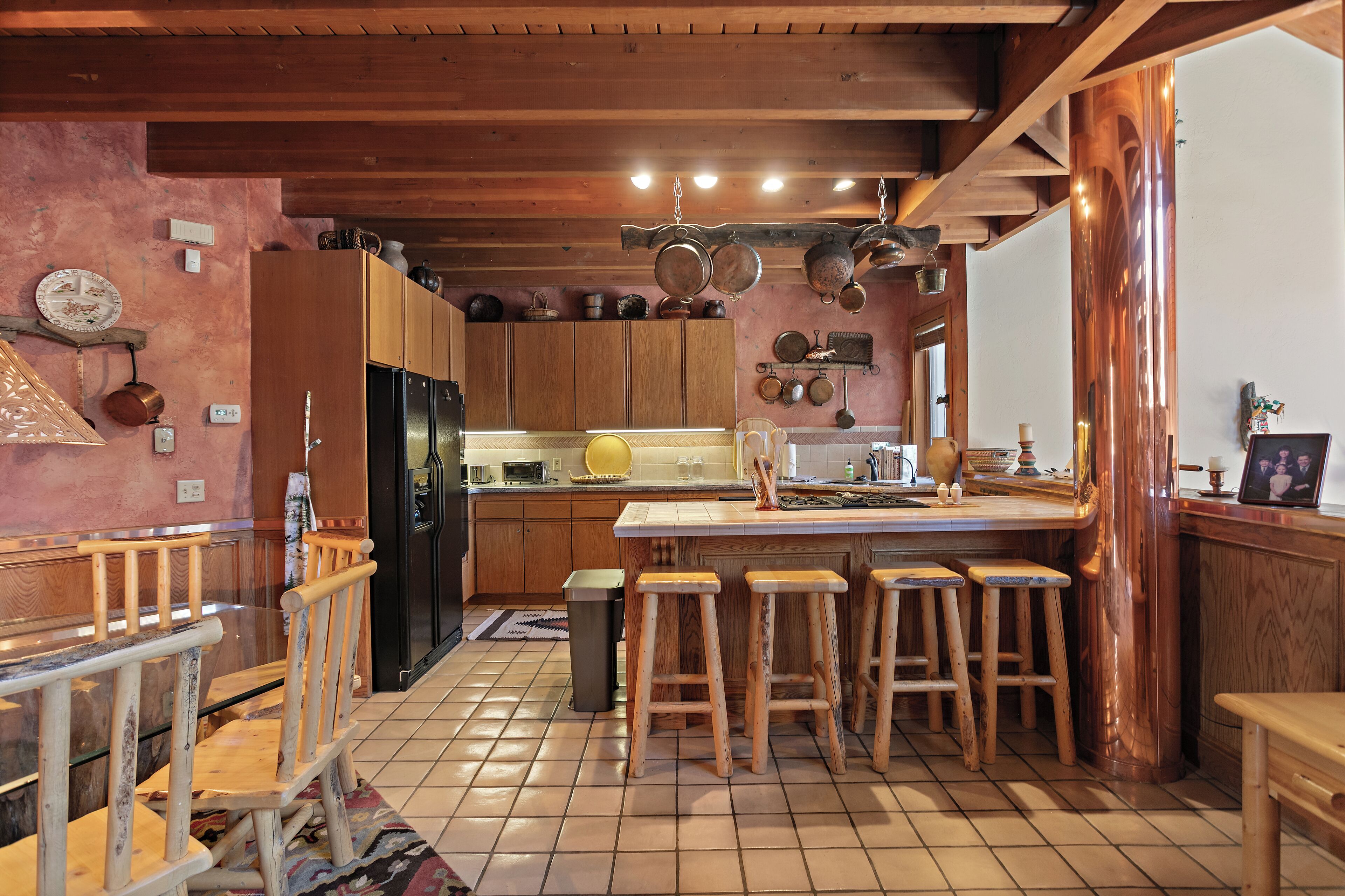 A kitchen with wooden cabinets, a central island with stools, hanging pots, and a tiled floor.