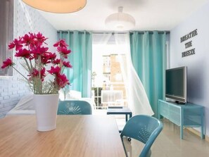 Ein modernes Wohnzimmer mit einem Holztisch, einem blauen Sessel, einer weißen Vase mit roten Blumen, einem Fernseher auf einem Schrank und einer Wand mit dem Schriftzug "BREATHE THE MUSIC".