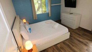 A bedroom with a bed, a TV, a window with curtains, a dresser, and a lamp. - Blue Sail Apartments - 1 bedroom apartments (sint maarten)