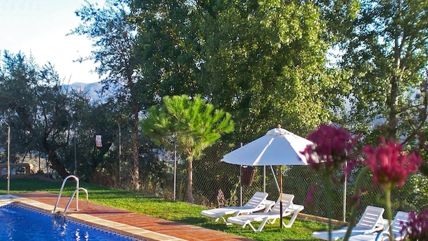 Una piscina con borde alicatado, rodeada de césped, árboles y una cerca. Se observan sillas de descanso y una sombrilla blanca.
