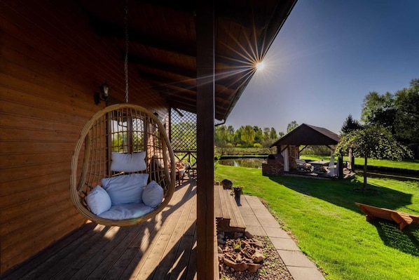 Una terraza de madera con una hamaca colgante, un espacio de estar cubierto y vistas a un estanque y jardín.