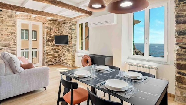 Una stanza accogliente con parete in pietra, un divano, un tavolo da pranzo per quattro persone e vista sul mare attraverso una finestra.