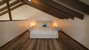 Un dormitorio con techo inclinado, una cama con ropa de cama blanca, dos lámparas de noche y una almohada azul.