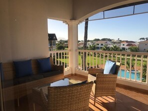 Ein Balkon mit Korbmöbeln, einem Glastisch und Blick auf ein Schwimmbecken und Häuser.