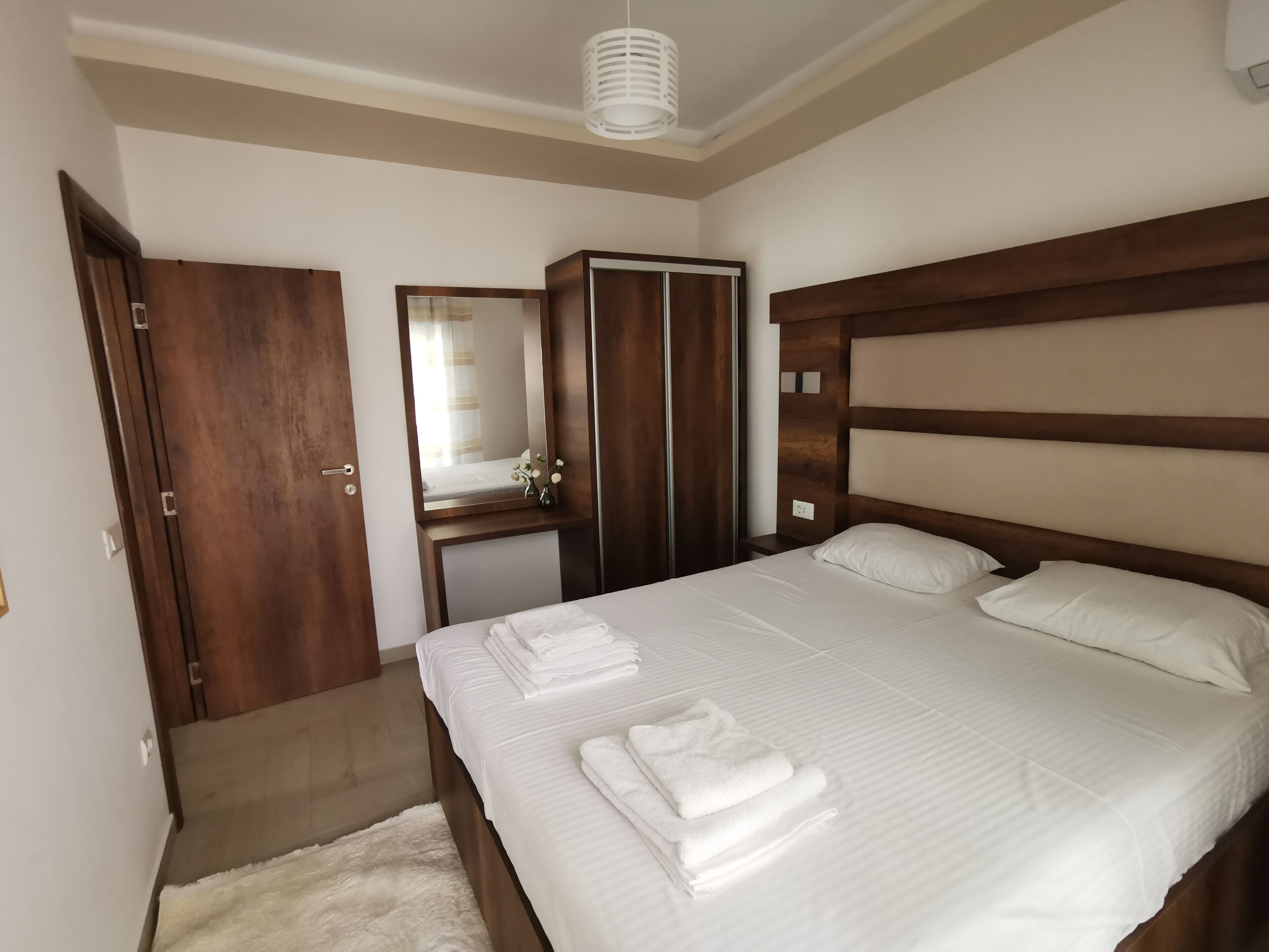 A neatly arranged bedroom with a large bed, a wooden wardrobe, a mirror, and a small bedside table.
