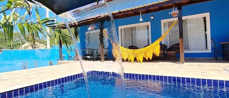 A swimming pool with a water feature, a hammock, and a building in the background.