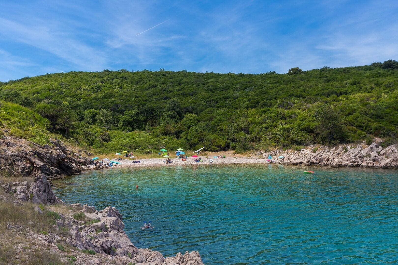 A coastal area with a rocky shoreline, clear turquoise water, and a densely forested hill in the background.