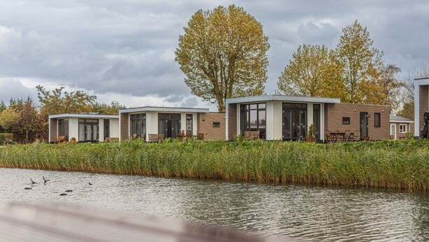 A row of modern, two-story houses with large windows and a flat roof, situated by a calm body of water with ducks visible.