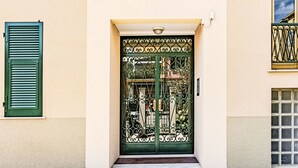L'ingresso di un edificio con un cancello in metallo verde, un piccolo gradino che conduce alla porta e una finestra con una griglia metallica sulla destra.