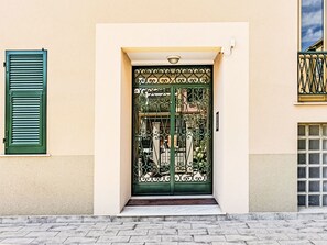 Ein Gebäudeeingang mit grünem Metallgitter, einem kleinen Podest vor der Tür und einem Fenster mit Metallgitter rechts.