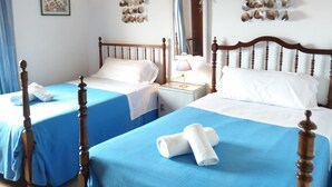 Habitación doble con dos camas, cada una con cabecero de madera, colcha azul y almohadas blancas. Hay una mesita de noche con una lámpara entre las camas, y un estante con caracolas sobre el espejo.