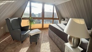 Un dormitorio con un ventanal grande, una cama, una silla, una mesita pequeña y una lámpara.