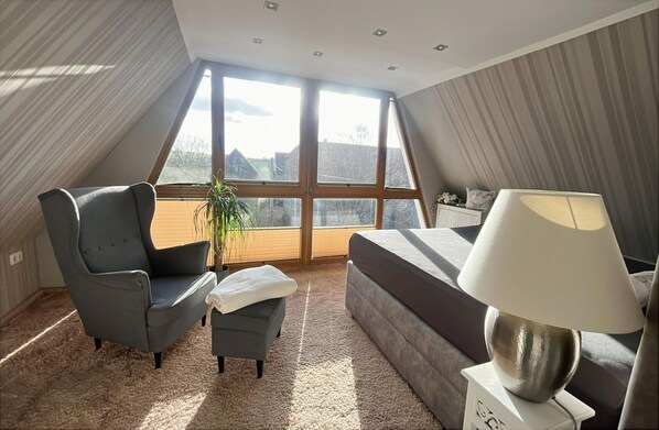 A bedroom with a large window, a bed, a chair, a small table, and a lamp. - Lieblingsplatz Appartement (Seelitz)
