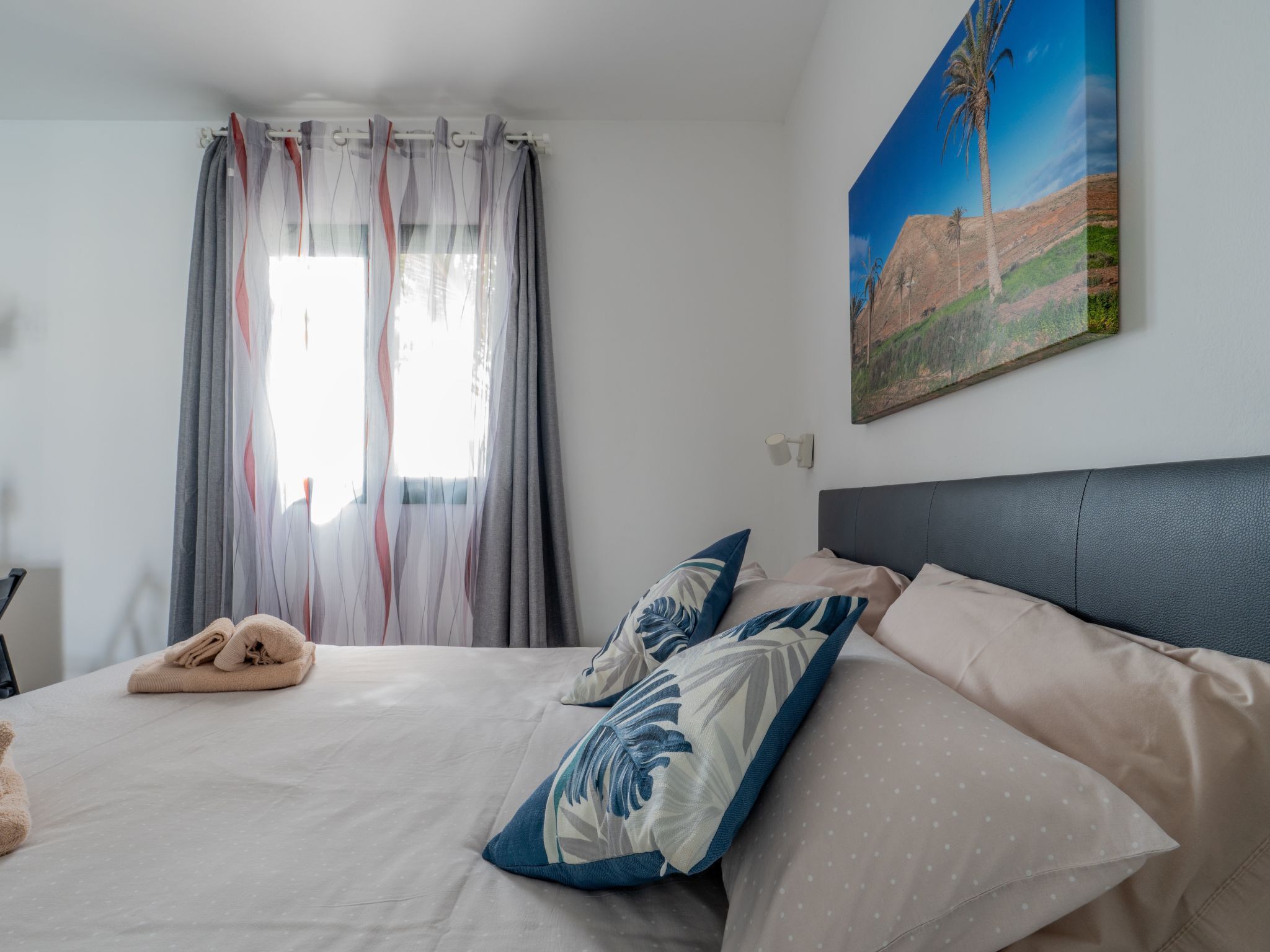 A neatly made bed with a pillow and a folded towel, a window with sheer curtains, and a painting of a landscape on the wall.