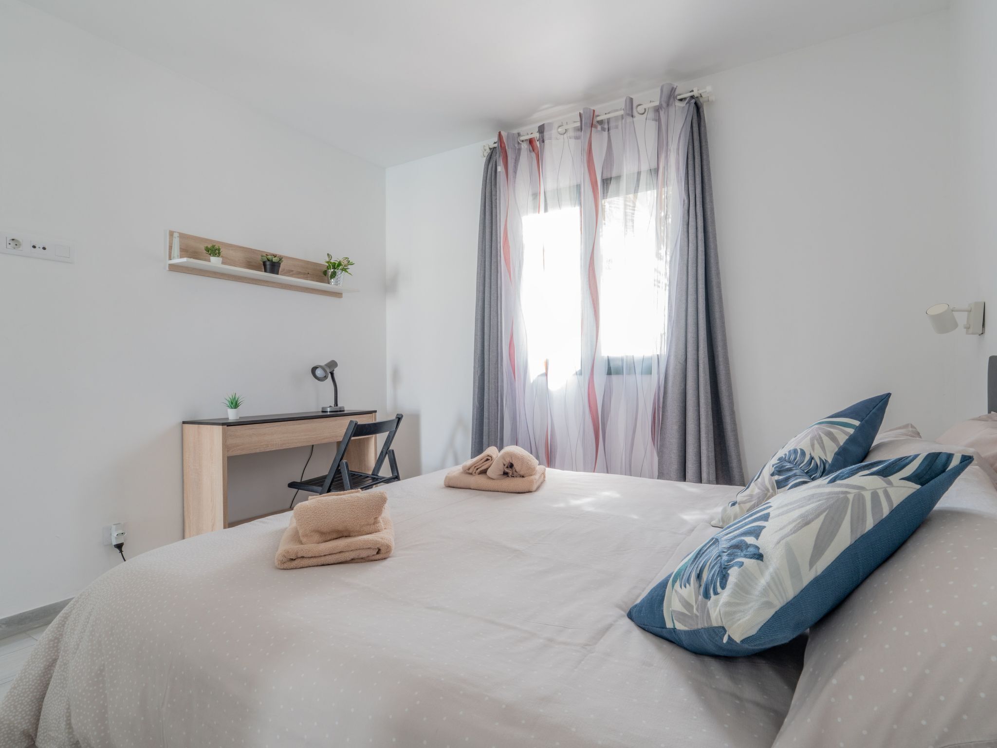 Een slaapkamer met een bed, een bureau en een stoel. Er is een raam met gordijnen, een plank met planten en een stopcontact.
