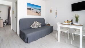 Un salon moderne avec un canapé gris, une table à manger entourée de chaises blanches, un tableau représentant une plage et un téléviseur à écran plat fixé au mur.