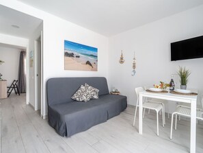 Un salon moderne avec un canapé gris, une table à manger entourée de chaises blanches, un tableau représentant une plage et un téléviseur à écran plat fixé au mur.